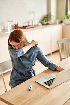 A woman sneezing at home, working on a laptop with a thermometer nearby.